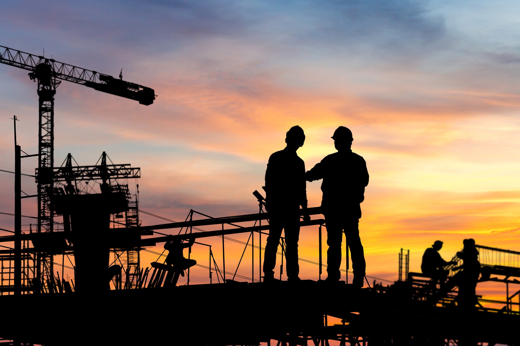 Construction workers silhouetted at a job site during sunset with cranes and industrial equipment